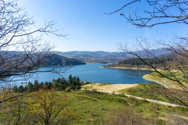 Alget rezervuarının bir parçası. Dalgalı koyu mavi su yüzeyi. Alçak kayalık kıyılar. Ön planda ağaç dalları. Orman, dağlar, arka planda mavi gökyüzü