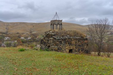 Ruins of the Church of St. Barbara (Barbare) of Sarkineti. Stone walls, collapsed bell tower dome. Trees without leaves, autumn grass. Cloudy sky and roofs of village houses in the background clipart