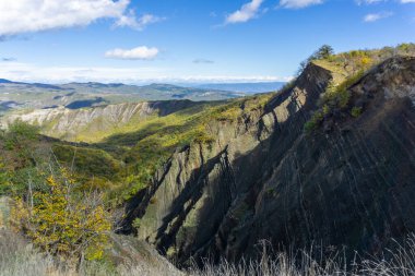 Çıplak kayalar bir vadiye iner. Jeoloji ve kaya katmanları görülebilir. Dağlar, tepeler ve arkasında bulutlar olan bir gökyüzü.