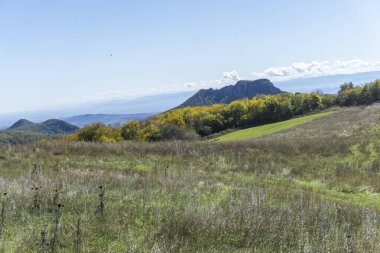 Kuru çimenli tarla, yeşil çayır, sonbahar yapraklı ağaçlar. Dağlar, tepeler ve gökyüzü.