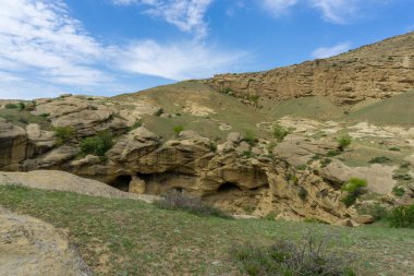 Kum taşı kayaları, çalılar ve taze otlarla kaplı taşlar. Arka planda bulutlar ve kayalar olan mavi gökyüzü
