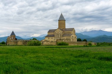 Kale duvarı, çan kulesi, konut binaları, Alaetti Manastırı Aziz George Kilisesi 'nin genel manzarası. Yeşil alan, bulutlu gökyüzü. Arka planda Kafkas Aralığı