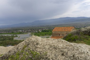 Eski bir şehrin tepesinde kiremitli çatısı olan kilise. Mağara kalıntıları. Arka planda vadi, nehir, köy ve dağlar var. Uplistsikhe eski bir kaya yontulmuş kasabadır..