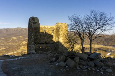 Jvari Manastırı 'nın sıcak akşam güneşiyle aydınlatılmış kale duvarı. Mavi gökyüzü, yapraksız ağaç, kocaman kayalar.