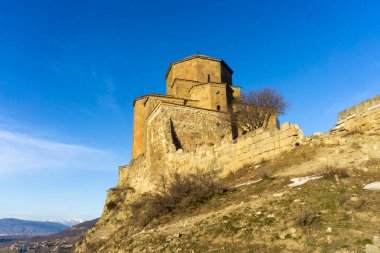 Jvari Manastırı sıcak akşam güneşiyle aydınlanır. Mavi gökyüzü, arka planda Kafkasya dağ sırasının bir parçası.