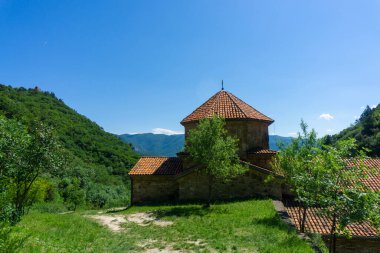 Church with orange tile roof of Shio-Mgvime monastery. Bright blue sky, rock, mountains with trees and bushes in the background clipart