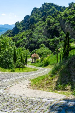 Shio-Mgvime Manastırı 'nın asfalt yolu dağın eteklerine iniyor. Ağaçlı tepeler, arka planda parlak mavi gökyüzü