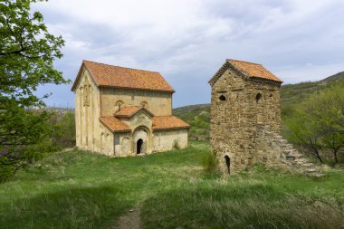 Maghlaant kilisesinin avlusunda kiremit çatılı ve taş kulesi olan taş kilise. Yeşil çimenler. Dağlar ve gökyüzü arkaplanda bulutlarla