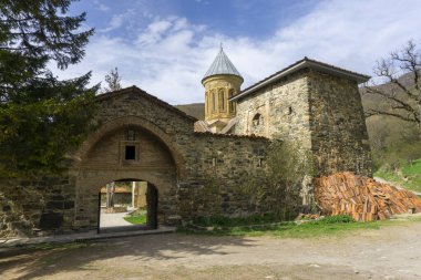 Kvatakhevi manastırının avlusuna giriş. Taş duvarlar, metal kapılar, kemer, kule, kilise kubbesi. Bulutlu parlak mavi gökyüzü.