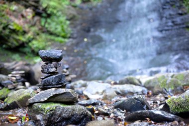 Huzurlu bir ormanda, bir dağ şelalesinin önünde dengeli bir taş yığını.