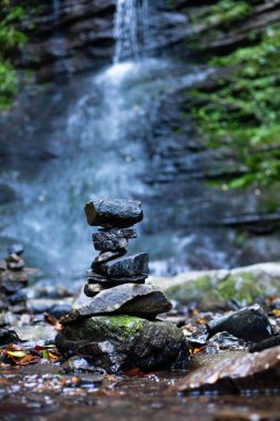 Huzurlu bir ormanda, bir dağ şelalesinin önünde dengeli bir taş yığını.