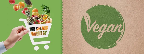 Vegan food online grocery shopping: hand holding a shopping cart icon full of vegetables and fruits