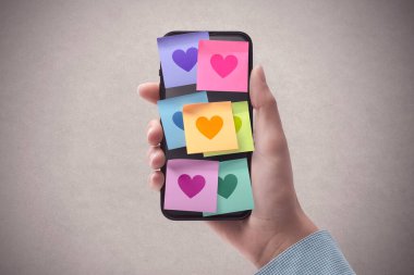 Woman holding a smartphone and lots of sticky notes with hearts on the display, relationships and communication concept, POV shot