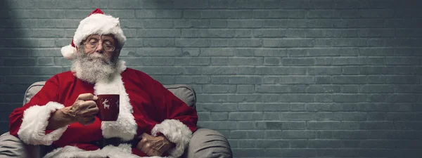 サンタクロースはアームチェアで家でリラックスし 熱い飲み物を持っています — ストック写真