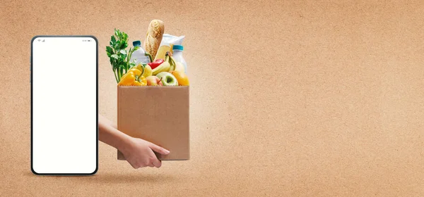 Online grocery and delivery service: smartphone with blank screen and hands holding a box full of fresh groceries