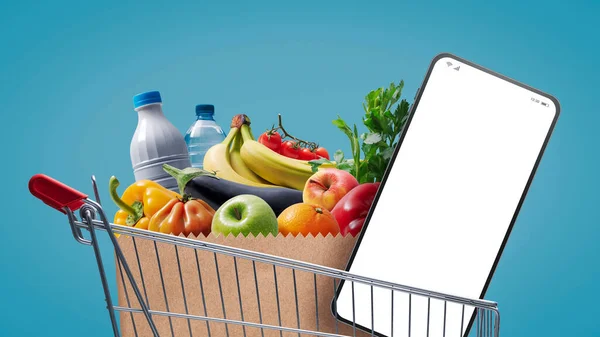 Supermarket shopping cart full of groceries and smartphone with blank screen, online grocery shopping concept
