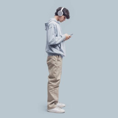 Cool teenager wearing headphones and connecting with his smartphone, isolated on gray background