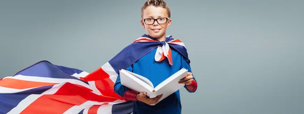 Cute superhero boy reading a book, he is wearing a British flag as a cape, education and enjoyment concept