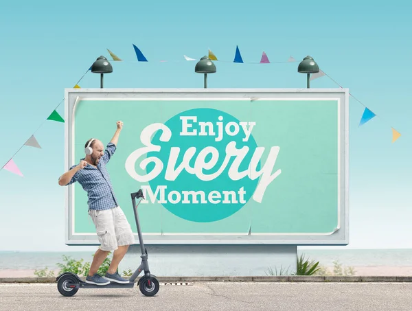 快乐的男人一边听着音乐 一边骑着一辆电子滑板车 背景是一张激励人的海报 享受每一个时刻 — 图库照片
