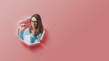 Yırtık kağıt deliğinden çıkıp elinde kitap, öğrenme ve eğitim konseptiyle gelen mutlu genç kız öğrenci.