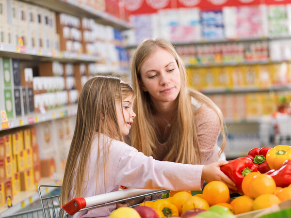Mother and daughter buying groceries together at the supermarket, the girl is picking vegetables in the produce section