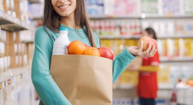 Elinde alışveriş poşeti olan gülümseyen genç bir kadın kameraya bakıyor, tezgahtar çalışıyor ve arka planda süpermarket rafları var.