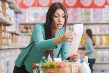 Süpermarkette şok olmuş genç bir kadın, bir alışveriş arabasına yaslanıyor ve uzun ve pahalı bir market fişini kontrol ediyor.