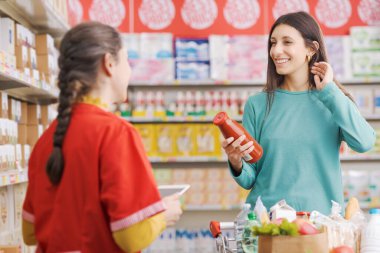 Dost canlısı bir market çalışanı bir kadına yardım ediyor, müşteri gülümsüyor ve elinde bir ürün tutuyor.