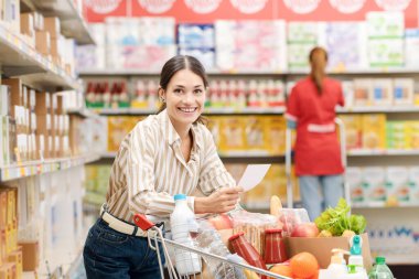 Marketten alışveriş yapan gülümseyen bir kadının portresi elinde bir alışveriş listesi ve dolu bir alışveriş arabası tutuyor.