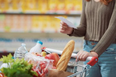 Markette market alışverişi yapan genç bir kadın, dolu bir tramvayı itiyor ve elinde bir alışveriş listesi tutuyor, kapat.