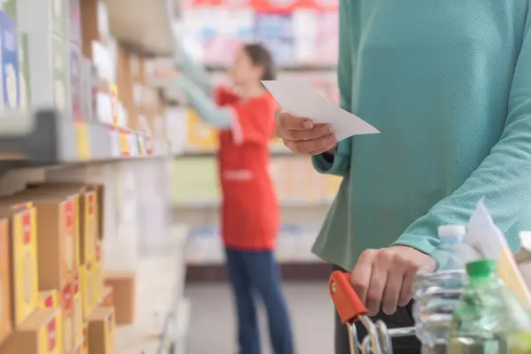 stock image Customer buying groceries at the supermarket, she is holding a grocery list and pushing a full shopping cart