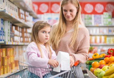 Annesiyle markette hayal kırıklığına uğramış bir kız elinde uzun ve pahalı bir market fişi tutuyor.