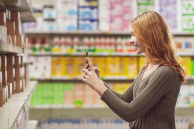 Markette market alışverişi yapan genç bir kadın akıllı telefonundaki bir yiyecek tarayıcı uygulamasını kullanarak yiyecek bilgilerini kontrol ediyor.