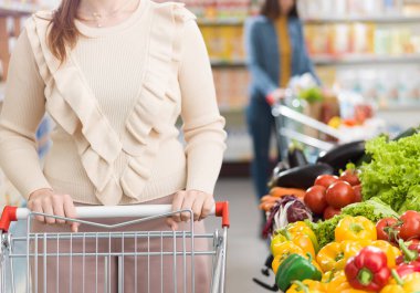 Süpermarketin sebze reyonunda taze sebzeler ve alışveriş arabasını iten bir kadın.