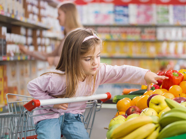Cute lovely girl sitting on the shopping cart, she is taking a fruit in the produce section while her mother is busy taking products on the shelves in the background