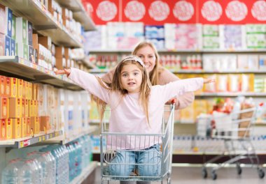 Markette alışveriş arabasında oturan neşeli tatlı kız, annesi onu zorluyor: market alışverişi ve yaşam tarzı konsepti.