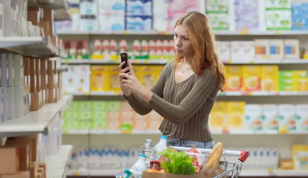 Young woman doing grocery shopping at the supermarket, she is checking food information using a food scanner app on her smartphone