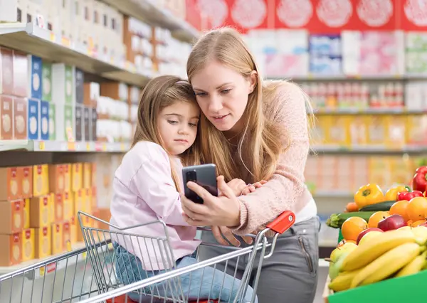 Cute girl sitting on a shopping cart at the supermarket, her mother is showing her apps on the smartphone: technology and grocery shopping concept