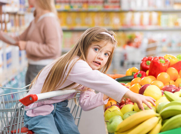 Cute surprised girl taking a fruit in the produce section at the supermarket, she is sitting on the shopping cart, healthy food concept