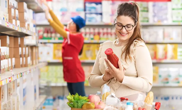 Woman doing grocery shopping at the supermarket and checking a food label on a product, stock clerk working in the background