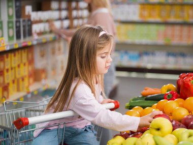 Alışveriş arabasında oturan sevimli kız, sebze reyonunda meyve toplarken annesi arka plandaki raflardaki ürünleri almakla meşgul.