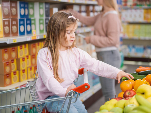 Cute lovely girl sitting on the shopping cart, she is taking a fruit in the produce section while her mother is busy taking products on the shelves in the background