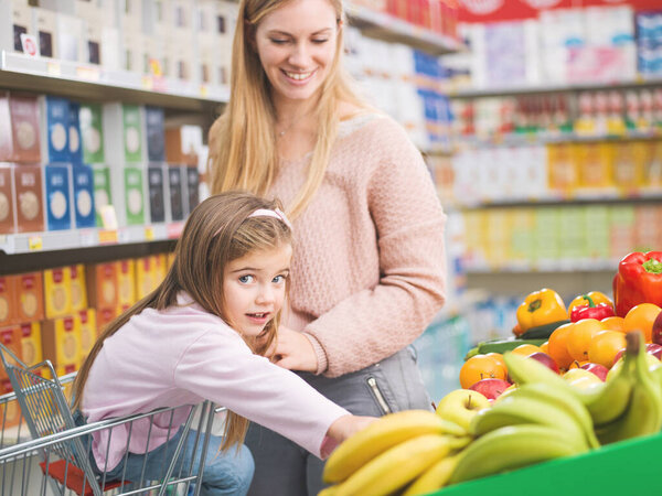 Happy cute girl sitting on a shopping cart and buying fresh vegetables and fruits with her mother at the supermarket