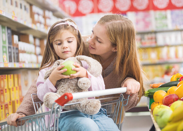 Mother and daughter doing grocery shopping together, the girl is holding a fruit and feeding her plushie