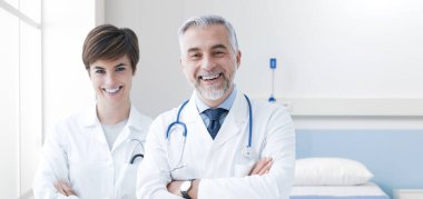 Kameraya poz veren ve gülümseyen kendinden emin doktorlar, arka planda hastane odası.