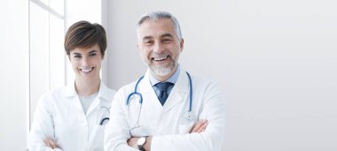 Kameraya poz veren ve gülümseyen kendinden emin doktorlar, arka planda hastane odası.