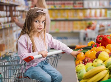 Süpermarkette meyve reyonunda meyve alan tatlı, şaşkın bir kız, alışveriş arabasında oturuyor, sağlıklı gıda konsepti.