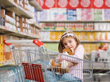 Süpermarkette gülümseyen tatlı bir kız, alışveriş arabasında oturmuş oyuncak ayısına sarılıyor.