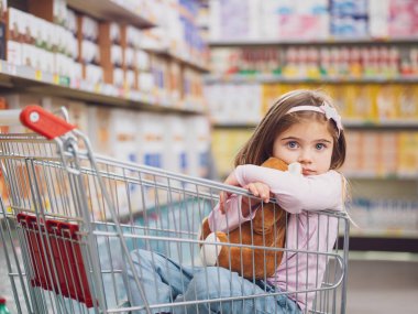 Süpermarkette küçük şirin bir kızın portresi. Bir alışveriş arabasının içinde oturmuş oyuncak ayısına sarılıyor.