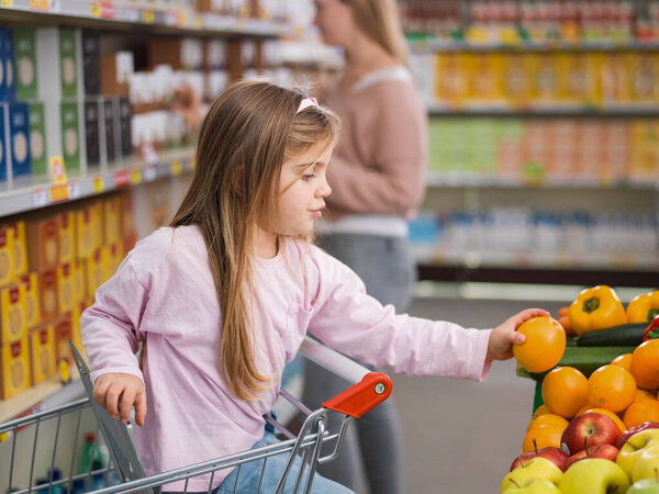 Smart girl sitting on the shopping cart and taking fresh healthy fruits at the supermarket, her mother is standing in the background